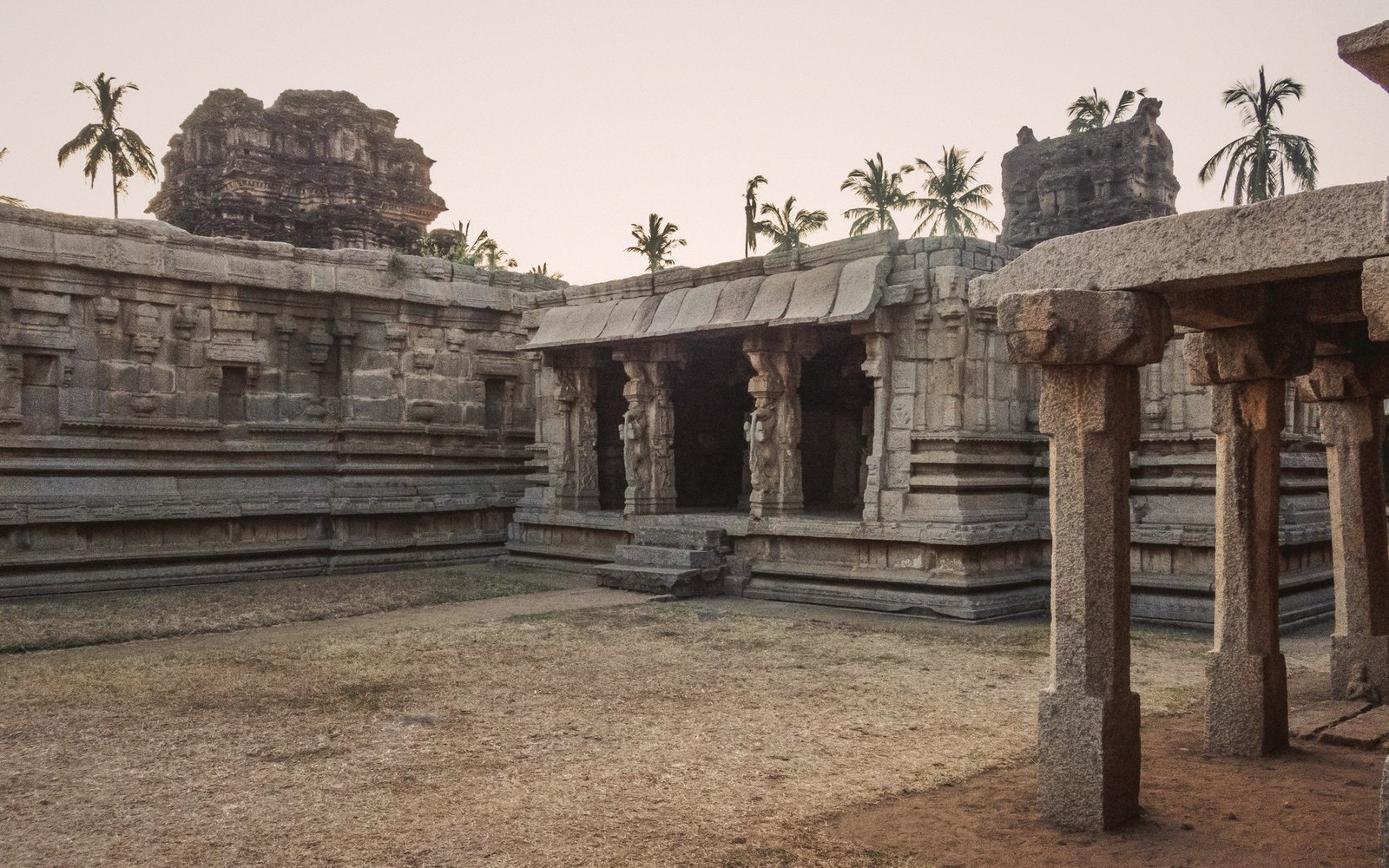 Ruined stone temple courtyard with carved pillars and crumbling gopuram towers behind