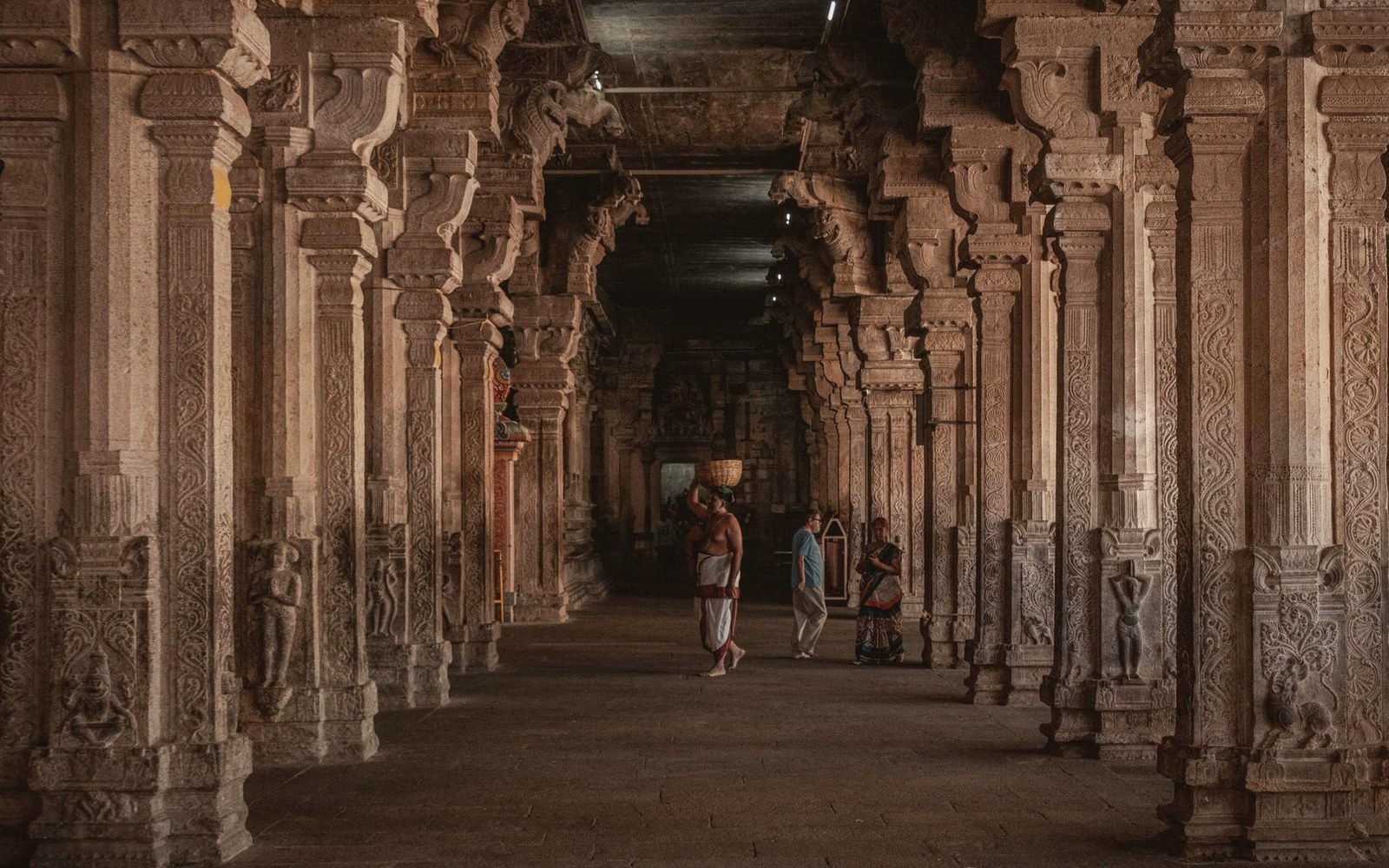 Carved stone pillars line a long temple corridor as visitors walk through